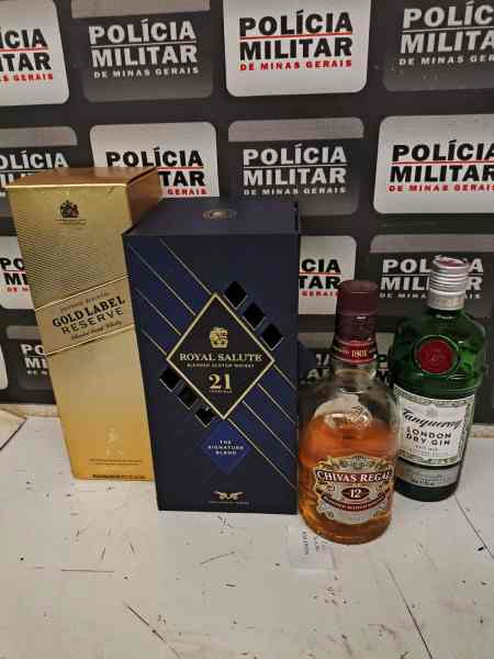 Garrafas de bebidas alcoólicas, incluindo Johnnie Walker Gold Label Reserve, Royal Salute e Chivas Regal, dispostas sobre uma superfície clara com fundo de cartaz da Polícia Militar.