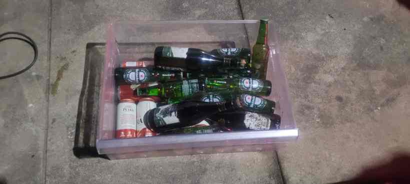 Caixa plástica transparente contendo várias garrafas de cerveja e refrigerantes, colocada sobre uma superfície de cimento.