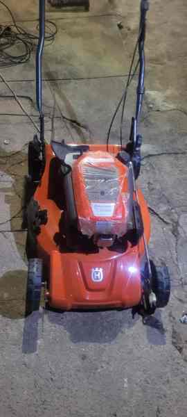 Uma máquina de cortar grama da marca Husqvarna, em cor vermelha, com proteção plástica sobre a parte superior, posicionada sobre uma superfície de piso áspero.