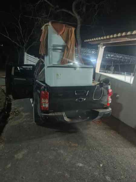 Caminhonete S10 preta com um freezer e um aparelho de refrigerador na carroceria, parada em um caminho de terra à noite.