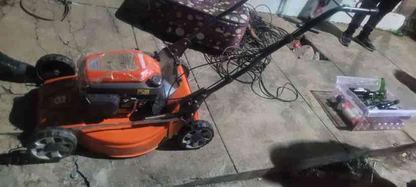 Uma máquina de cortar grama laranja, localizada em um piso de concreto, com uma caixa plástica transparente cheia de garrafas ao fundo.