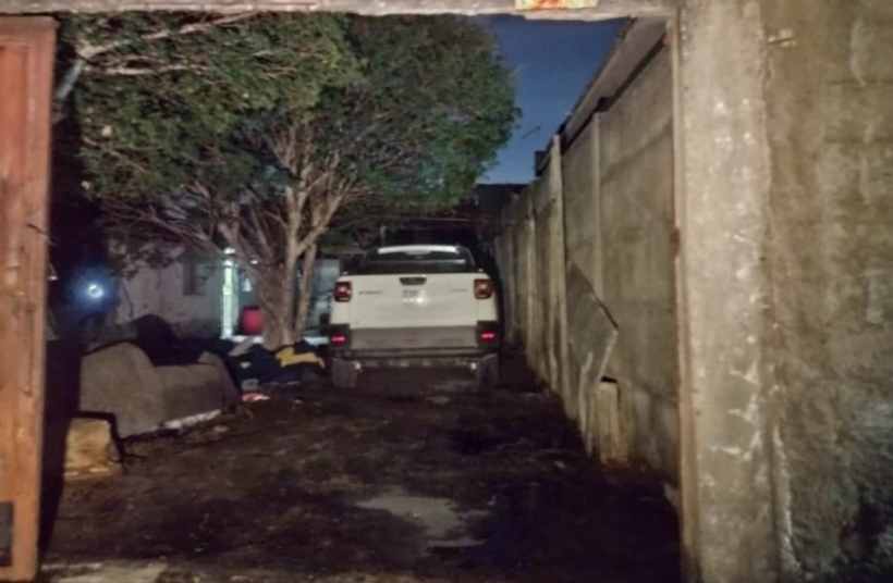 Imagem de um quintal escuro, onde uma caminhonete Fiat Strada branca está estacionada, visível através de um portão. O ambiente é cercado por muros e árvores.