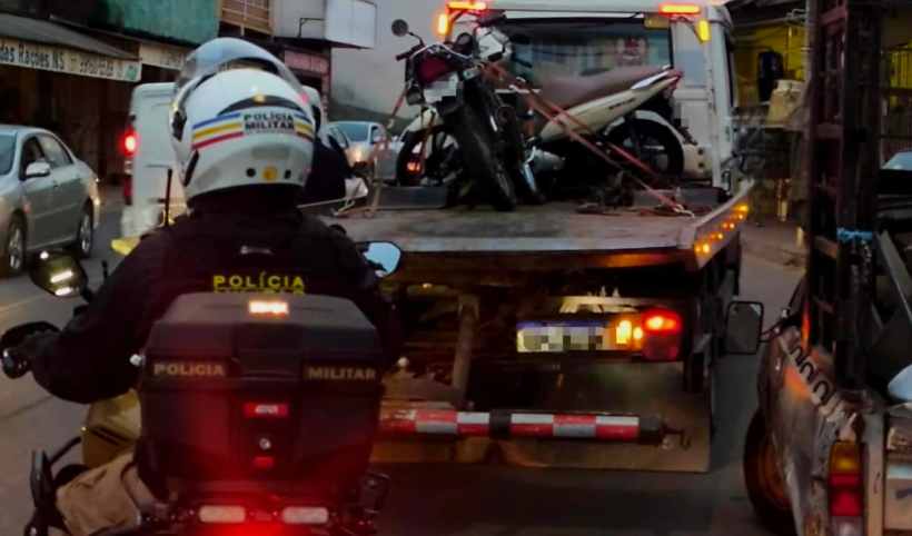Motocicleta sendo rebocada por um caminhão, com um policial militar em uma motocicleta observando a ação durante a Operação Cavalo de Aço em Nova Serrana.