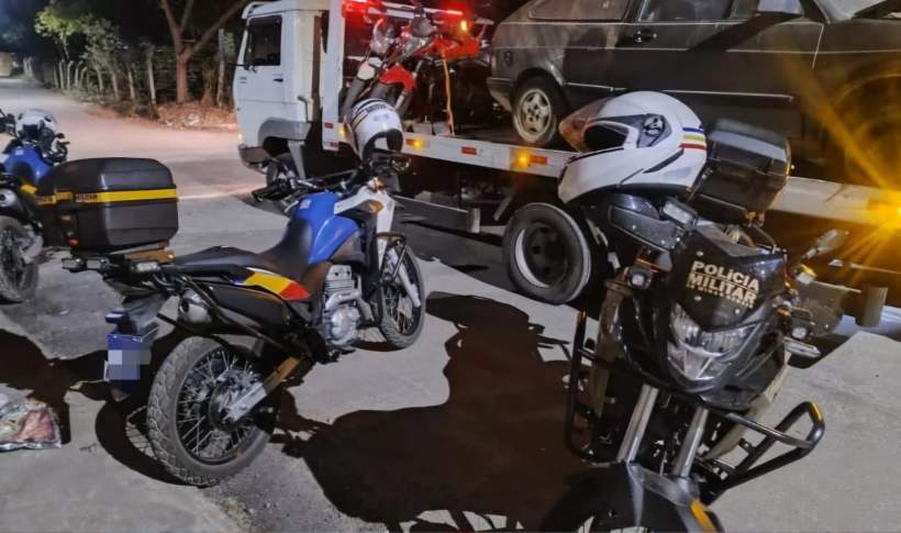 Imagem de motos da Polícia Militar de Minas Gerais estacionadas próximas a um caminhão guincho, com um veículo sendo carregado. A cena ocorre em uma rua à noite.