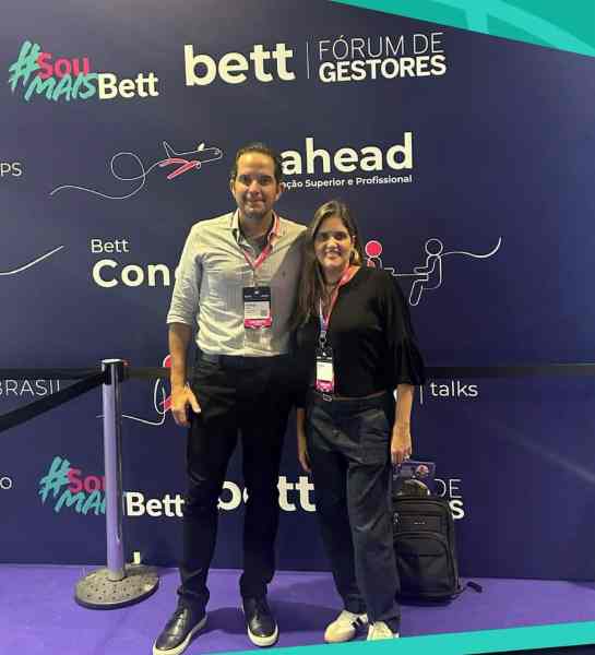 Duas pessoas posando para uma foto em frente a um fundo com as palavras 'Bett', 'Fórum de Gestores' e 'ahead', em um evento educacional.