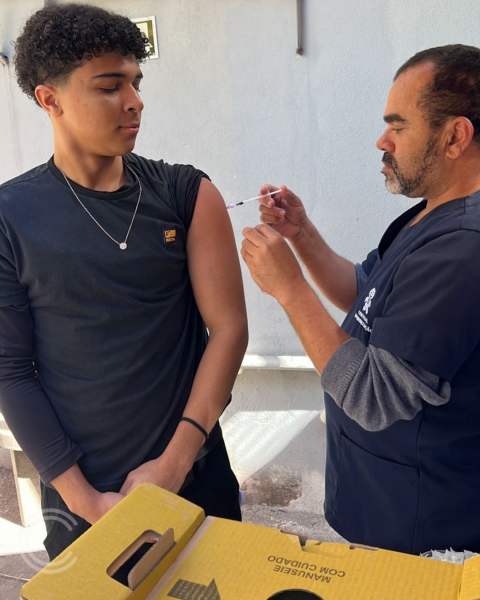 Um profissional de saúde administrando uma vacina em um jovem, em ambiente externo. O jovem está de camiseta preta e o profissional usa um uniforme de saúde.