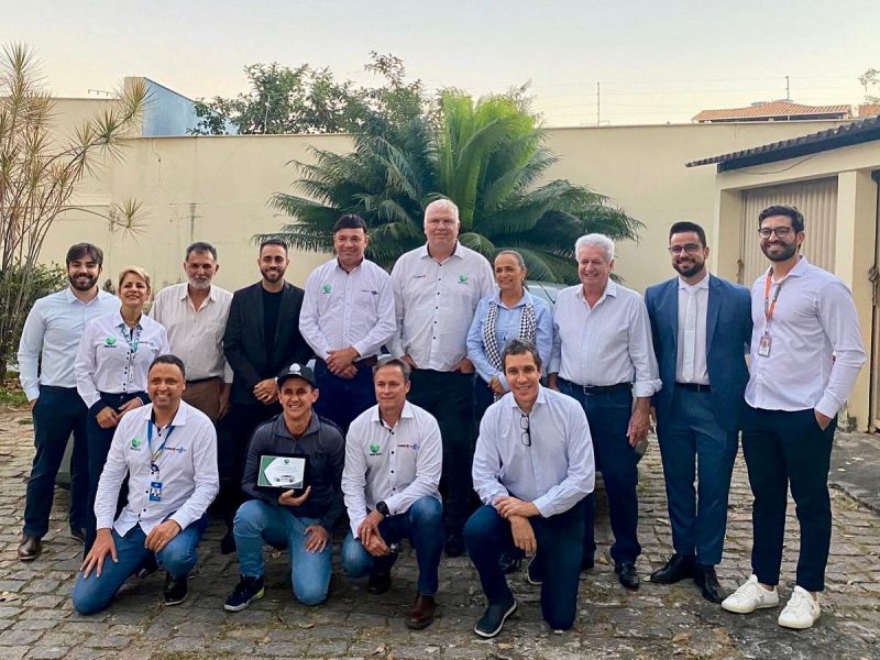 Grupo de integrantes da Agência Novo Oeste posando para foto após a entrega de um veículo novo, com um fundo de vegetação e um edifício.
