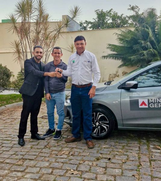 Três homens em frente a um veículo novo, um deles entregando as chaves do carro, com um ambiente de arbustos e vegetação ao fundo.