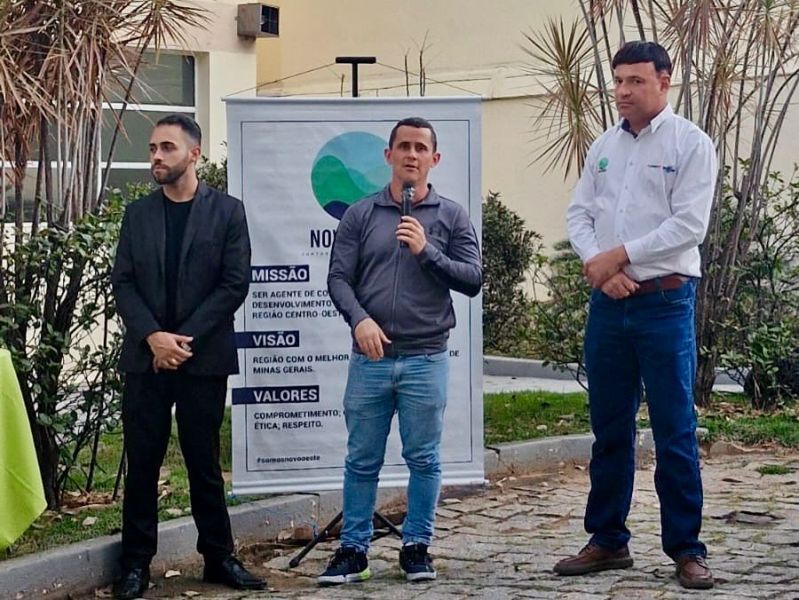 Três homens em pé, um falando ao microfone, em evento da Agência Novo Oeste, com cartaz ao fundo destacando a missão e valores da agência.