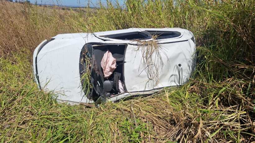 Veículo VW Gol capotado em um campo coberto de grama alta após saída de pista, com partes do interior visíveis.
