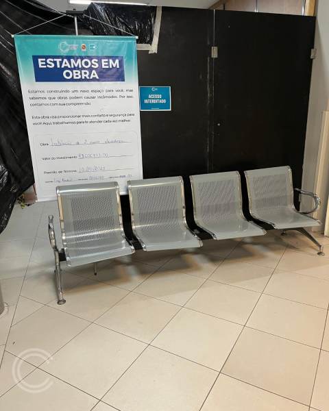 Bancos de espera de metal prateado organizados em uma sala de espera, com um aviso na parede indicando que a área está em obra.