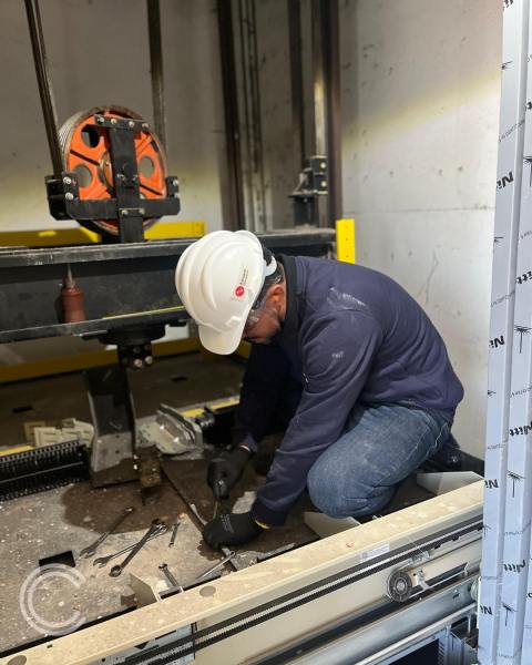 Técnico trabalhando na instalação de um elevador, usando capacete de segurança e ajustando componentes no interior do poço do elevador.