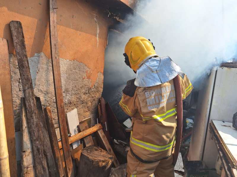 Um bombeiro em traje de combate a incêndio, com capacete, está lutando contra as chamas em uma cozinha ao ar livre, cercado por materiais inflamáveis e fumaça.