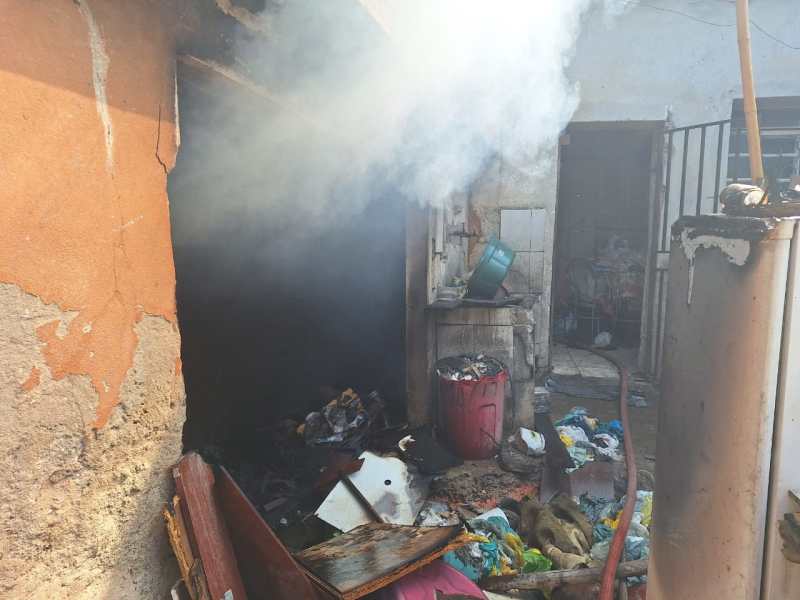Cenário de incêndio em uma cozinha externa, com fumaça saindo do interior e restos de materiais queimados ao redor.
