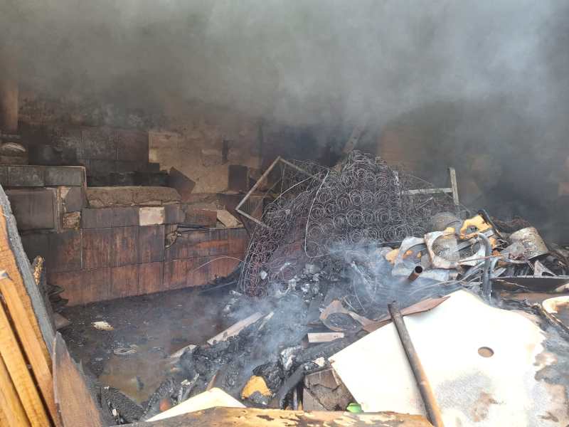 Interior de uma residência com fumaça densa e material queimado após incêndio, incluindo caixas de madeira e uma estrutura de metal, em um ambiente desordenado.