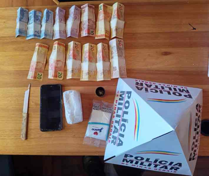 Imagem de uma mesa com dinheiro em notas de diferentes valores, um celular, um pacote de drogas embrulhado em papel e um recipiente pequeno, além de uma faca ao lado. Também é visível uma caixa com a identificação da Polícia Militar.