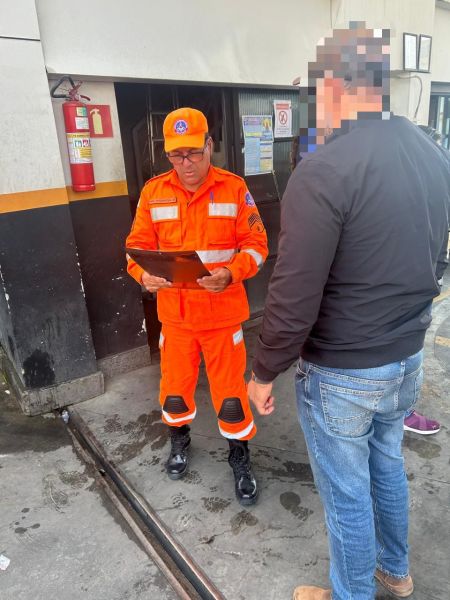 Agente do Corpo de Bombeiros Militar em uniforme laranja conversando com um homem em um posto de combustíveis durante uma fiscalização.