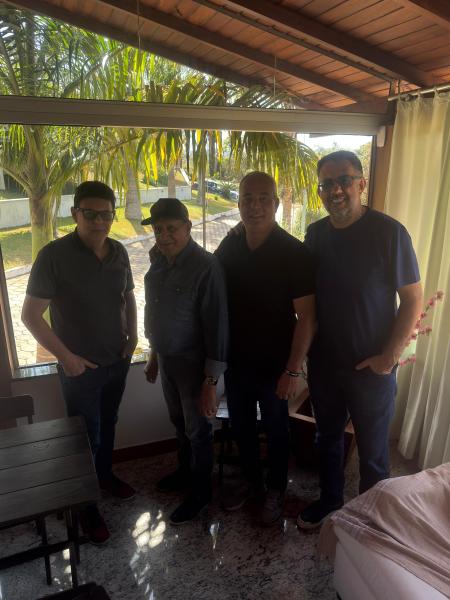 Quatro homens posando juntos em um ambiente interno, com luz natural entrando pela janela e palmeiras visíveis do lado de fora.