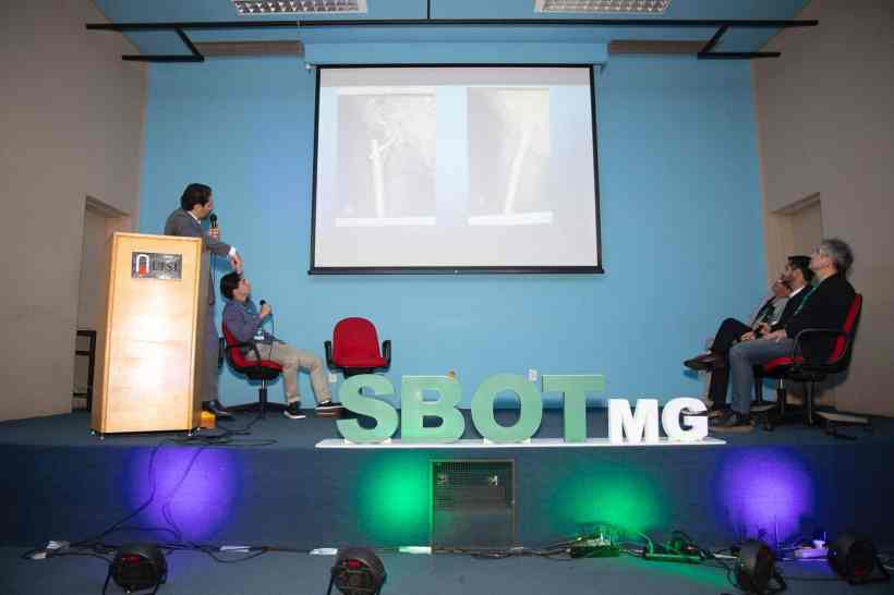 Apresentação durante o curso da SBOT-MG, com palestrante mostrando uma imagem em um projetor, e participantes assistindo em cadeiras no palco.