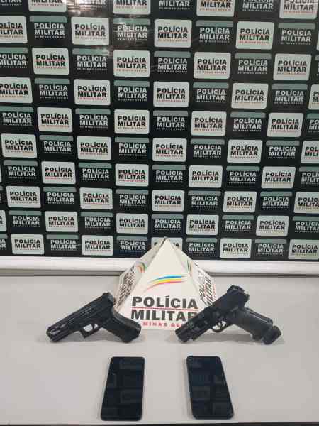 Duas pistolas e dois celulares sobre uma mesa, com um fundo que exibe repetidamente o logo da Polícia Militar.