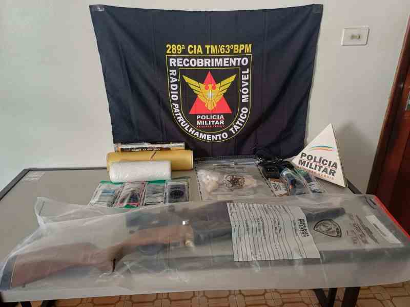 Materiais apreendidos durante operação da Polícia Militar, incluindo armas, munições, drogas e equipamentos, com uma bandeira da polícia ao fundo.
