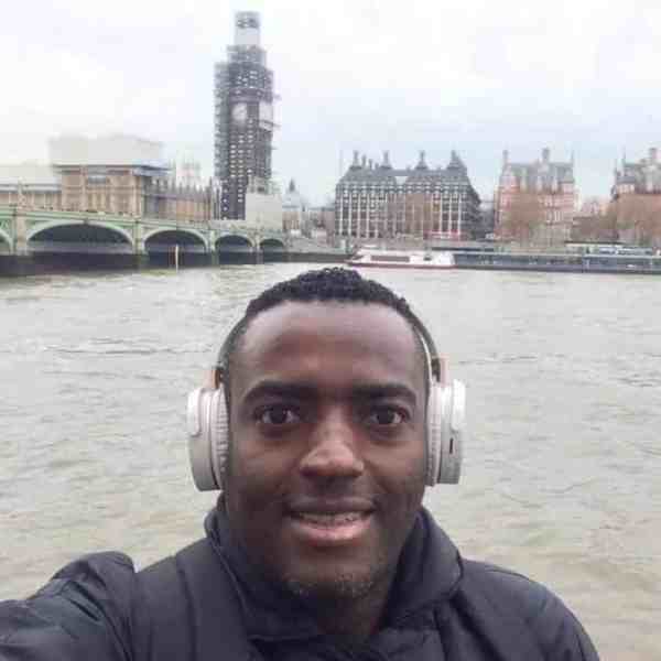 Selfie de um homem usando fones de ouvido, posando em frente ao rio com edifícios famosos ao fundo, incluindo uma construção em reforma.