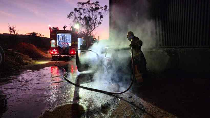Bombeiro combatendo incêndio em veículo com mangueira, próximo a um caminhão bomba, ao amanhecer.