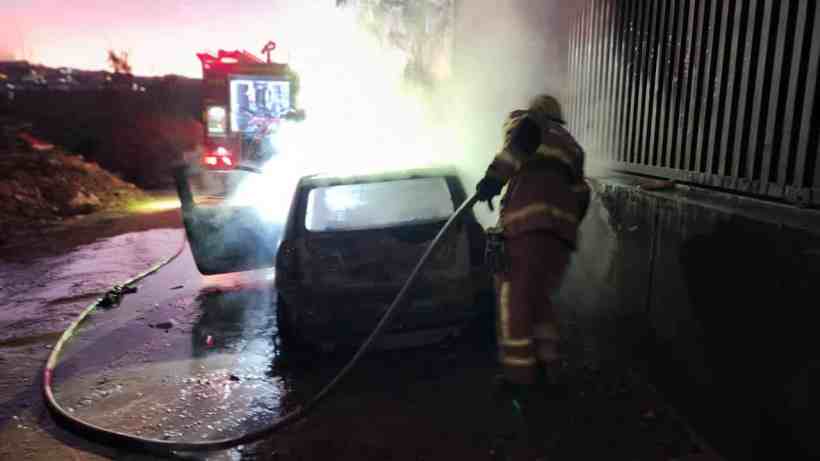 Bombeiro combatendo incêndio em veículo em Nova Serrana, com fumaça saindo do carro e caminhão de bombeiros ao fundo.