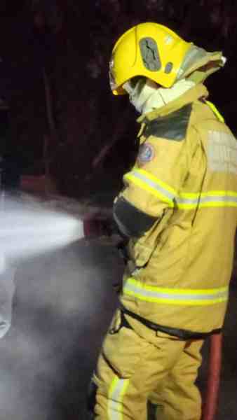 Bombeiro apagando um incêndio em veículo à noite, vestindo uniforme completo e capacete amarelo.