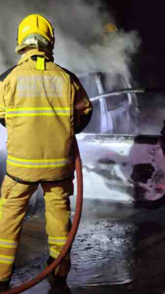 Bombeiro combatendo incêndio em veículo, com fumaça e chamas visíveis, em ambiente noturno.