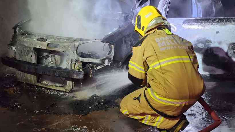 Bombeiro combatendo incêndio em veículo danificado, com fumaça visível ao fundo.