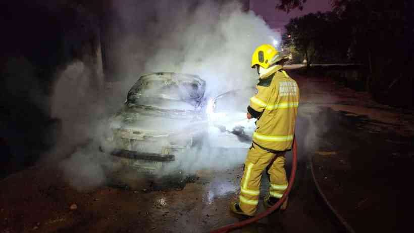 Bombeiro combatendo incêndio em veículo, rodeado por fumaça e com mangueira na mão.