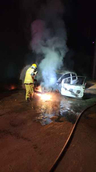 Bombeiro combatendo incêndio em veículo, com fumaça e chamas visíveis, durante a noite.