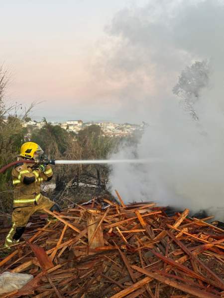 Bombeiro combatendo incêndio em madeireira, com mangueira, diante de pilhas de madeira em chamas e fumaça.