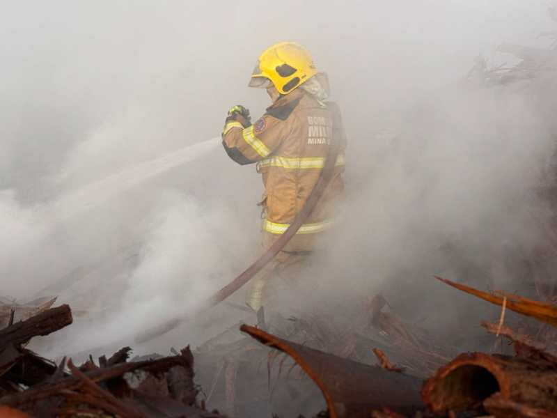 Um bombeiro do Corpo de Bombeiros Militar de Minas Gerais combate um incêndio em uma madeireira, cercado por fumaça e restos de madeira.