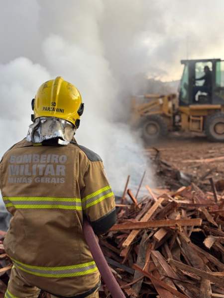 Bombeiro militar em ação, usando mangueira para combater incêndio em madeireira, com fumaça visível e maquinário ao fundo.