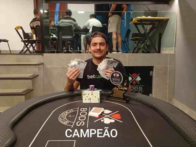 Um jovem sorridente segurando várias notas de dinheiro, celebrando sua vitória em um torneio de poker, sentado em uma mesa de poker com um troféu e cartas visíveis ao seu redor.