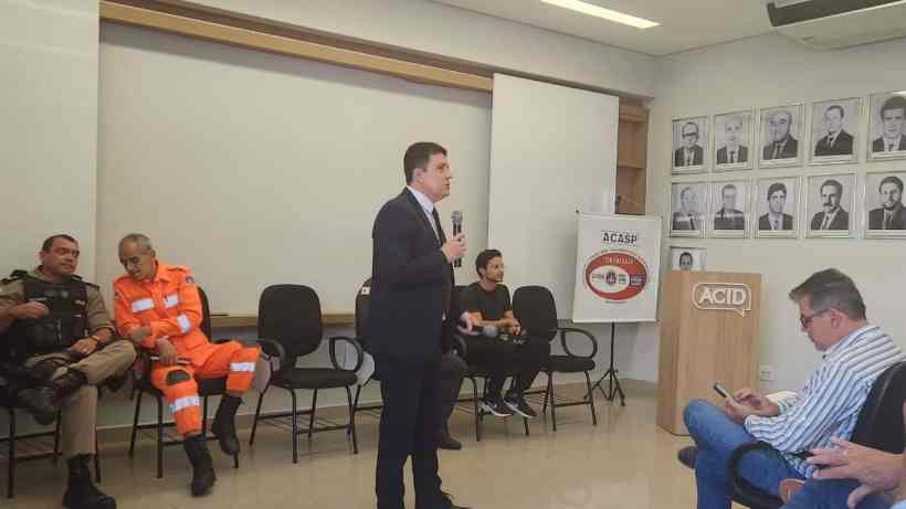Reunião da ACASP no auditório da ACID, com representantes da sociedade civil e autoridades discutindo segurança pública, mobilidade e políticas de acolhimento social. Figura mostra um palestrante em pé, falando ao microfone, enquanto outros participantes ouvem e estão sentados em cadeiras.