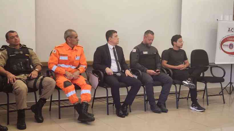 Cinco homens sentados em cadeiras em um auditório, próximo a uma mesa. Eles estão vestidos com uniformes de segurança, incluindo um oficial da polícia, um bombeiro, e outros em roupas civis. O ambiente é formal, com um banner da ACASP ao fundo.