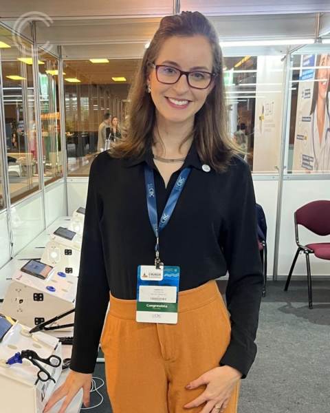 Dra. Isabella Alves Almeida Machado posando em um evento médico, vestindo uma camisa preta e calças largas, com um crachá de identificação e equipamentos cirúrgicos ao fundo.