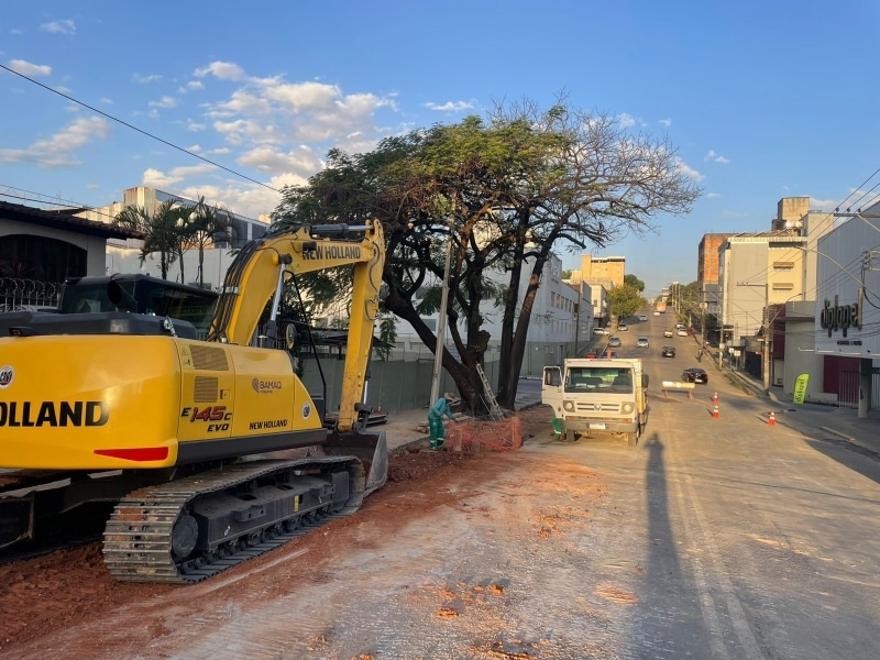 Máquina pesada de escavação amarela em uma rua em obras, com árvores e edificações ao fundo.