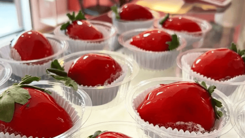 Vários morangos do amor cobertos com gelatina vermelha, apresentados em copinhos descartáveis, com folhas verdes visíveis.