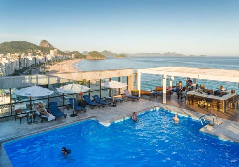 Piscina em um hotel no Rio de Janeiro, com vista para a praia e montanhas ao fundo, cercada por espreguiçadeiras e guarda-sóis.