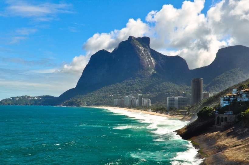 Vista da praia com ondas do mar e montanha ao fundo em um dia ensolarado no Rio de Janeiro.