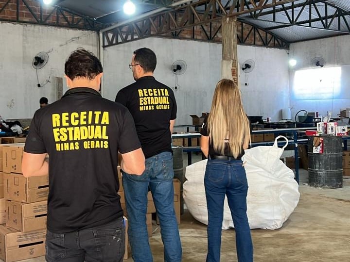 Agentes da Receita Estadual de Minas Gerais inspecionando uma fábrica clandestina de sabão em pó falsificado em um galpão, com caixas de produtos ao fundo.