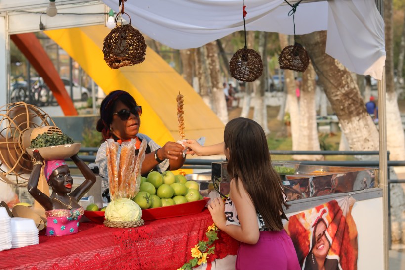 Uma mulher vende um espetinho para uma garota em um estande de festival, com frutas e vegetais decorando a mesa.