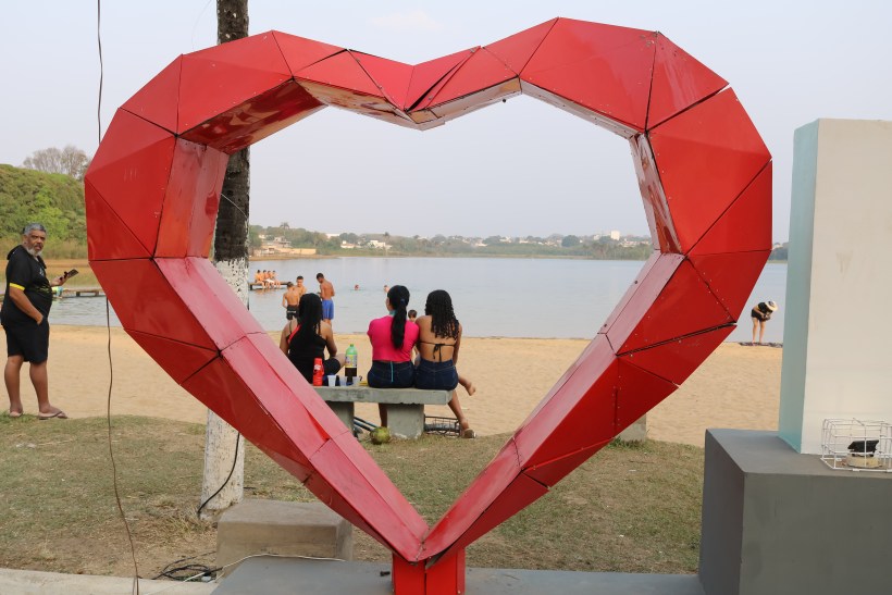 Escultura em forma de coração vermelho em um espaço à beira da água, com pessoas sentadas em um banco ao fundo e outras se divertindo na água.