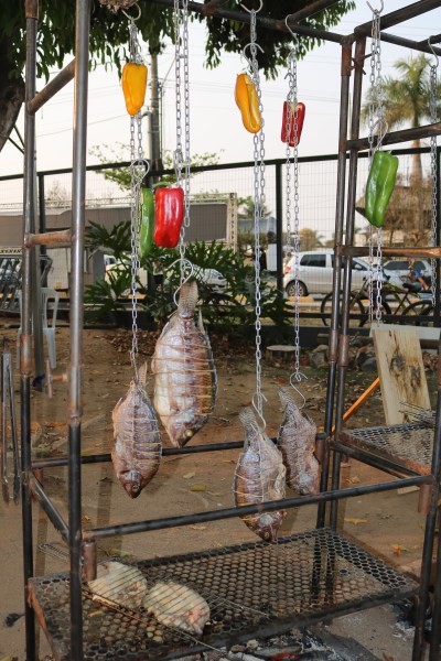 Peixes sendo grelhados em churrasqueira com pimentões coloridos pendurados, ambientado em um espaço ao ar livre.