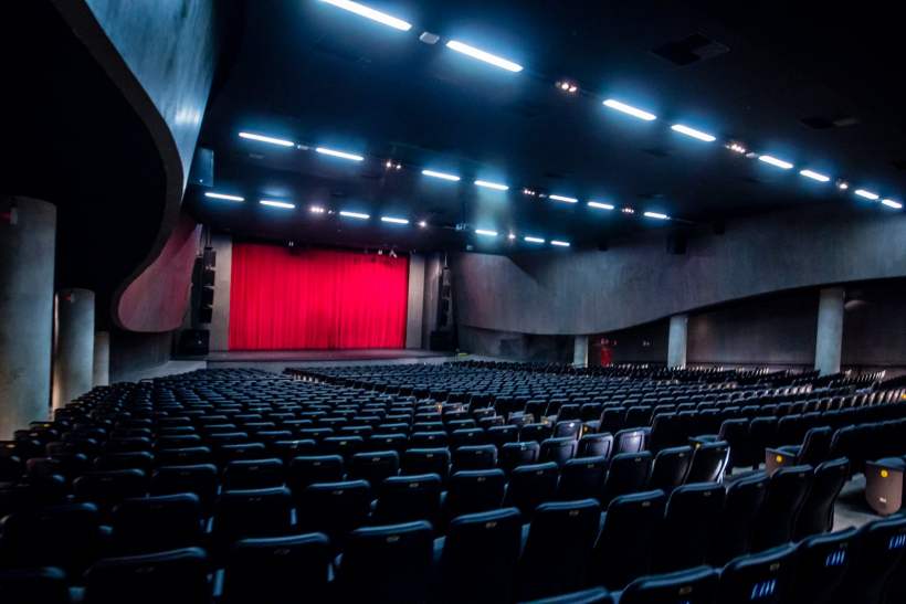 Interior de um teatro moderno, com luzes LED no teto e um palco iluminado por cortinas vermelhas, com assentos vazios em primeiro plano.