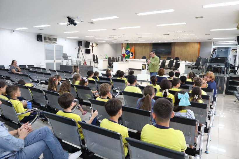 Um grupo de crianças em camisetas amarelas participa de uma palestra em um auditório, com uma mesa e bandeiras ao fundo. O palestrante fala para os alunos que estão sentados em fileiras.